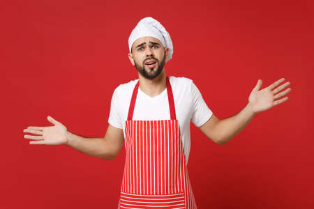 Perplexed young bearded male chef cook or baker man in striped apron white t-shirt toque chefs hat posing isolated on bright red background. Cooking food concept. Mock up copy space. Spreading hands.の写真素材