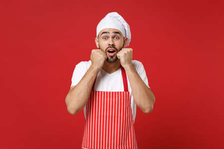 Shocked young bearded male chef cook or baker man in striped apron white t-shirt toque chefs hat posing isolated on bright red background. Cooking food concept. Mock up copy space. Clenching fists.の写真素材