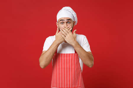 Shocked young bearded male chef cook or baker man in striped apron white t-shirt toque chefs hat posing isolated on red background. Cooking food concept. Mock up copy space. Covering mouth with hands.の写真素材