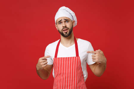Perplexed young male chef cook or baker man in striped apron white t-shirt toque chefs hat posing isolated on red background. Cooking food concept. Mock up copy space. Point index fingers on himself.の写真素材