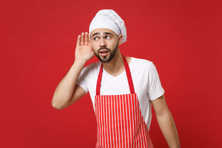 Curious bearded young male chef cook or baker man in striped apron white t-shirt toque chefs hat posing isolated on red background in studio. Cooking food concept. Mock up copy space. Try to hear you.の写真素材