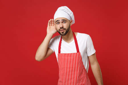 Bearded young male chef cook or baker man in striped apron white t-shirt toque chefs hat posing isolated on red background. Cooking food concept. Mock up copy space. Try to hear with hand near ear.の写真素材
