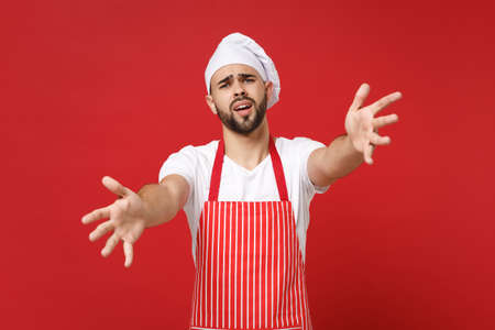 Displeased young male chef cook or baker man in striped apron white t-shirt toque chefs hat posing isolated on red background. Cooking food concept. Mock up copy space. Stand with outstretched hands.の写真素材