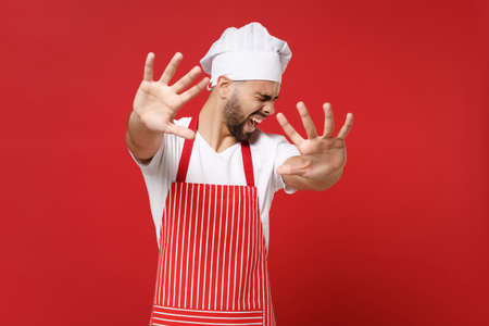 Disgusted scared young male chef cook or baker man in striped apron white t-shirt toque chefs hat posing isolated on red wall background. Cooking food concept. Mock up copy space. Covering with hands.の写真素材