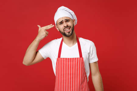 Unhappy young male chef cook baker man in striped apron toque chefs hat posing isolated on red background. Cooking food concept. Mock up copy space. Point fingers to head as if he about shoot himself.の写真素材