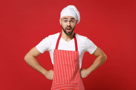 Shocked irritated young male chef cook baker man in striped apron white t-shirt toque chefs hat posing isolated on red background. Cooking food concept. Mock up copy space. Standing with arms akimbo.の写真素材