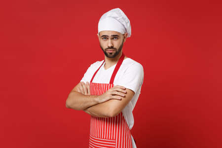 Handsome young bearded male chef cook baker man in striped apron white t-shirt toque chefs hat posing isolated on red wall background. Cooking food concept. Mock up copy space. Holding hands crossed.の写真素材