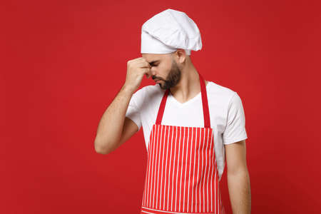 Young bearded male chef cook baker man in striped apron white t-shirt toque chefs hat posing isolated on red background. Cooking food concept. Mock up copy space. Keeping eyes closed put hand on nose.の写真素材