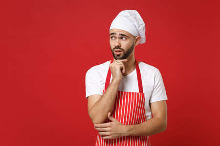 Pensive male chef cook baker man in striped apron white t-shirt toque chefs hat posing isolated on red background. Cooking food concept. Mock up copy space. Put hand prop up on chin, looking aside.の写真素材