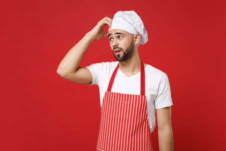 Pensive young male chef cook baker man in striped apron white t-shirt toque chefs hat posing isolated on red background. Cooking food concept. Mock up copy space. Putting hand on head, looking aside.の写真素材