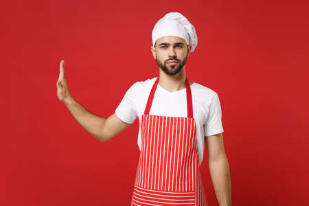 Serious young male chef cook baker man in striped apron white t-shirt toque chefs hat posing isolated on red background. Cooking food concept. Mock up copy space. Showing stop gesture aside with palm.の写真素材