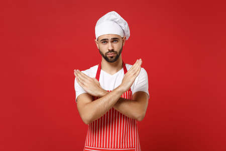Young male chef cook baker man in striped apron white t-shirt toque chefs hat posing isolated on red wall background. Cooking food concept. Mock up copy space. Showing stop gesture with crossed hands.の写真素材