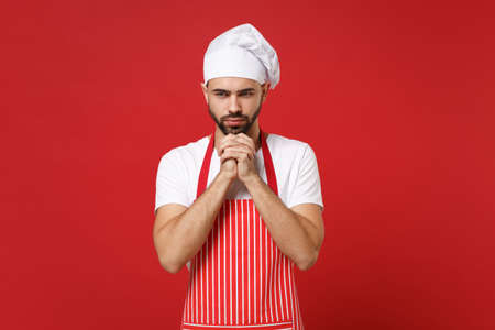 Pensive male chef cook baker man in striped apron white t-shirt toque chefs hat posing isolated on red background. Cooking food concept. Mock up copy space. Put hands prop up on chin, looking down.の写真素材