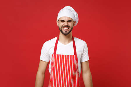 Angry irritated young bearded male chef cook baker man in striped apron white t-shirt toque chefs hat posing isolated on red wall background. Cooking food concept. Mock up copy space. Showing teeth.の写真素材
