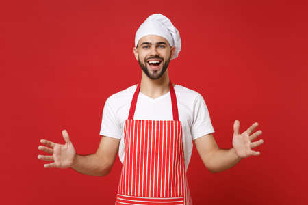 Cheerful young chef cook baker man in striped apron toque chefs hat posing isolated on red background. Cooking food concept. Mock up copy space. Gesturing demonstrating size with horizontal workspace.の写真素材