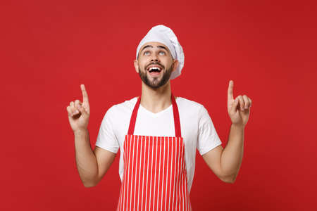Amazed young bearded male chef cook baker man in striped apron white t-shirt toque chefs hat posing isolated on red background. Cooking food concept. Mock up copy space. Pointing index fingers up.の写真素材
