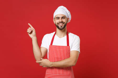 Smiling young bearded male chef cook baker man in striped apron white t-shirt toque chefs hat posing isolated on red background. Cooking food concept. Mock up copy space. Pointing index finger aside.の写真素材