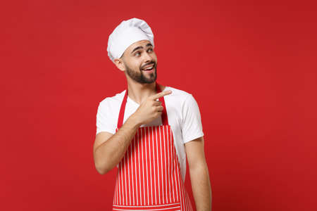 Handsome young bearded male chef cook baker man in striped apron white t-shirt toque chefs hat posing isolated on red background. Cooking food concept. Mock up copy space. Pointing index finger aside.の写真素材