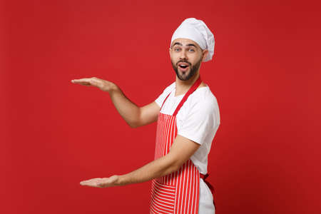 Excited young chef cook baker man in striped apron toque chefs hat posing isolated on red background. Cooking food concept. Mock up copy space. Gesturing demonstrating size with vertical workspace.の写真素材