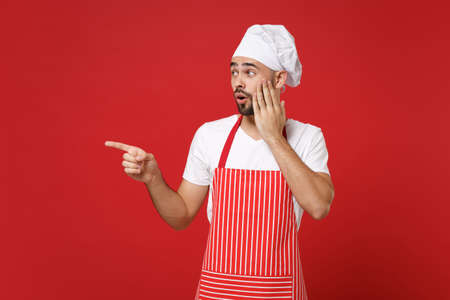 Shocked chef cook baker man in striped apron white t-shirt toque chefs hat posing isolated on red background. Cooking food concept. Mock up copy space. Pointing index fingers aside put hand on cheek.の写真素材