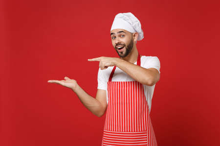 Cheerful young male chef cook baker man in striped apron white t-shirt toque chefs hat posing isolated on red background. Cooking food concept. Mock up copy space. Pointing index finger, hand aside.の写真素材