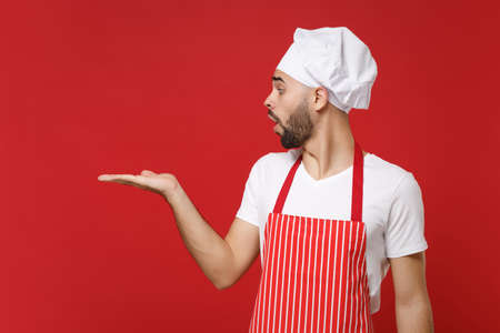 Shocked young male chef cook baker man in striped apron white t-shirt toque chefs hat posing isolated on red background. Cooking food concept. Mock up copy space. Pointing hand aside, hold something.の写真素材