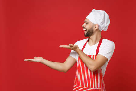 Laughing young bearded male chef cook baker man in striped apron white t-shirt toque chefs hat posing isolated on red wall background. Cooking food concept. Mock up copy space. Pointing hands aside.の写真素材