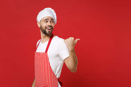 Side view of smiling young bearded chef cook baker man in striped apron white t-shirt toque chefs hat posing isolated on red background. Cooking food concept. Mock up copy space. Pointing thumb aside.の写真素材