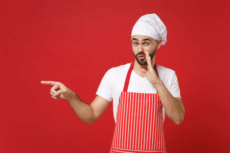 Secret chef cook baker man in apron toque chefs hat posing isolated on red background. Cooking food concept. Mock up copy space. Whispers gossip, tells secret with hand gesture, pointing finger aside.の写真素材