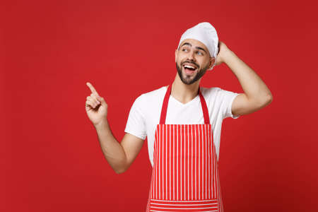Funny chef cook baker man in striped apron white t-shirt toque chefs hat posing isolated on red background. Cooking food concept. Mock up copy space. Putting hand on head, pointing index finger aside.の写真素材