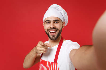 Close up of chef cook baker man in striped apron toque chefs hat posing isolated on red background. Cooking food concept. Mock up copy space. Doing selfie shot on mobile phone, point finger on camera.の写真素材