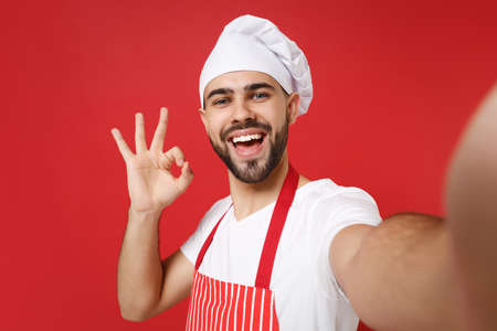 Close up of chef cook baker man in striped apron toque chefs hat posing isolated on red background. Cooking food concept. Mock up copy space. Doing selfie shot on mobile phone, showing OK gesture.の写真素材