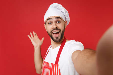 Close up of excited young bearded chef cook baker man in striped apron toque chefs hat posing isolated on red background. Cooking food concept. Mock up copy space. Doing selfie shot on mobile phone.の写真素材
