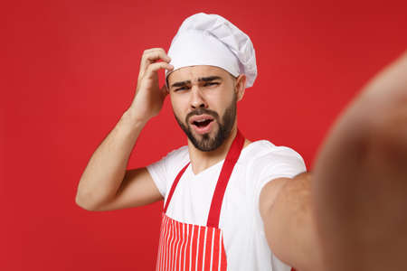 Close up of preoccupied bearded chef cook baker man in striped apron toque chefs hat posing isolated on red background. Cooking food concept. Mock up copy space. Doing selfie shot on mobile phone.の写真素材