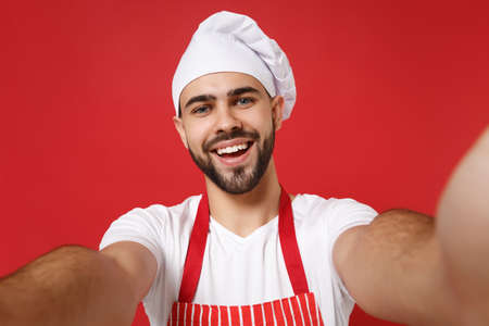 Close up of smiling young bearded chef cook baker man in striped apron toque chefs hat posing isolated on red background. Cooking food concept. Mock up copy space. Doing selfie shot on mobile phone.の写真素材