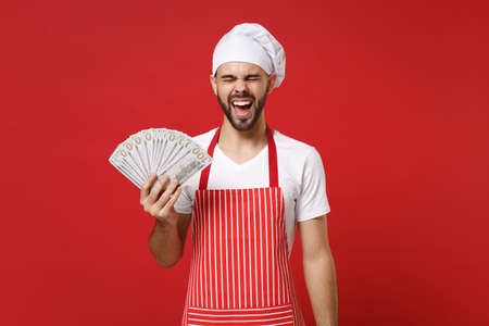 Screaming young bearded male chef cook or baker man in striped apron toque chefs hat isolated on red background. Cooking food concept. Mock up copy space. Hold fan of cash money in dollar banknotes.の写真素材