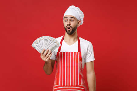 Amazed young bearded male chef cook or baker man in striped apron toque chefs hat isolated on red background. Cooking food concept. Mock up copy space. Holding fan of cash money in dollar banknotes.の写真素材