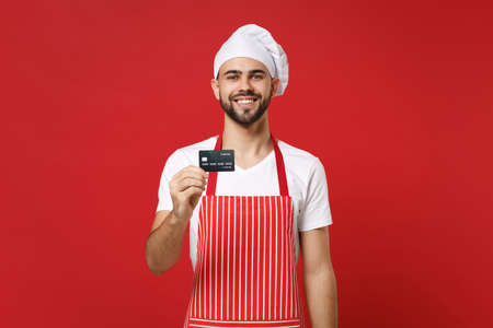 Smiling young bearded male chef cook or baker man in striped apron white t-shirt toque chefs hat isolated on red wall background. Cooking food concept. Mock up copy space. Holding credit bank card.の写真素材