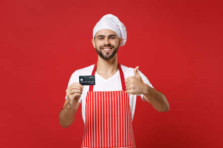Smiling young male chef cook or baker man in striped apron white t-shirt toque chefs hat isolated on red background. Cooking food concept. Mock up copy space. Hold credit bank card, showing thumb up.の写真素材