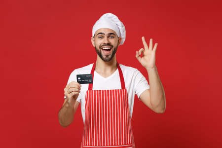 Joyful young male chef cook or baker man in striped apron white t-shirt toque chefs hat isolated on red background. Cooking food concept. Mock up copy space. Hold credit bank card, showing OK gesture.の写真素材