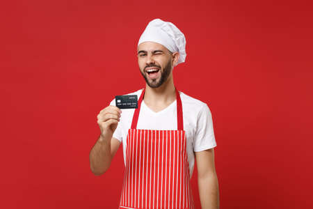 Blinking young bearded male chef cook or baker man in striped apron white t-shirt toque chefs hat isolated on red wall background. Cooking food concept. Mock up copy space. Holding credit bank card.の写真素材