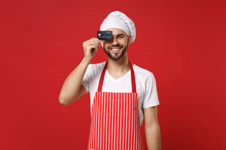 Funny young bearded male chef cook or baker man in striped apron white t-shirt toque chefs hat isolated on red background. Cooking food concept. Mock up copy space. Covering eye with credit bank card.の写真素材