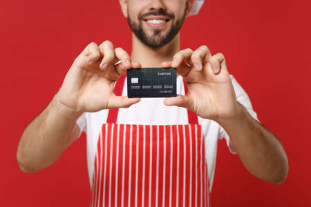Cropped image of young bearded male chef cook or baker man in striped apron white t-shirt isolated on bright red wall background. Cooking food concept. Mock up copy space. Holding credit bank card.の写真素材