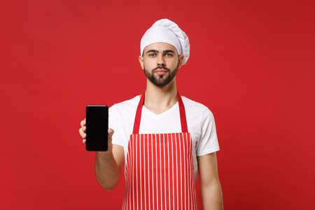 Serious young bearded male chef cook or baker man in striped apron toque chefs hat isolated on red background. Cooking food concept. Mock up copy space. Holding mobile phone with blank empty screen.の写真素材