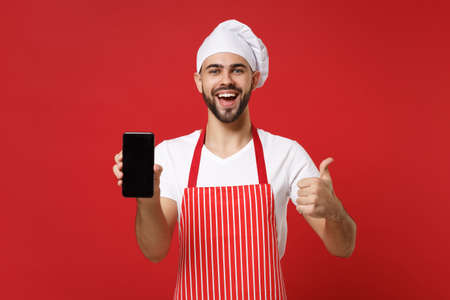 Cheerful male chef cook or baker man in striped apron toque chefs hat isolated on red background. Cooking food concept. Mock up copy space. Hold mobile phone with blank empty screen showing thumb up.の写真素材