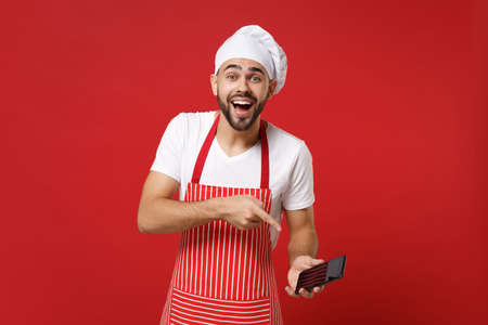 Laughing young bearded male chef cook or baker man in striped apron toque chefs hat isolated on bright red background. Cooking food concept. Mock up copy space. Pointing index finger on mobile phone.の写真素材