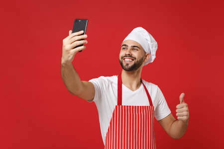 Smiling young chef cook or baker man in striped apron toque chefs hat isolated on red wall background. Cooking food concept. Mock up copy space. Doing selfie shot on mobile phone, showing thumb up.の写真素材