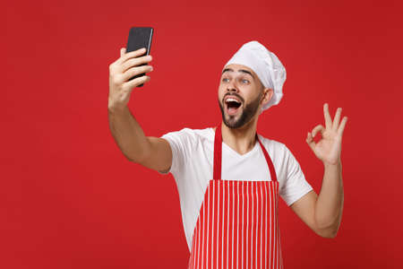 Excited young chef cook or baker man in striped apron toque chefs hat isolated on red wall background. Cooking food concept. Mock up copy space. Doing selfie shot on mobile phone, showing OK gesture.の写真素材