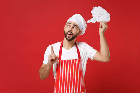 Pensive chef cook or baker man in striped apron toque chefs hat isolated on red background. Cooking food concept. Mock up copy space. Hold say cloud with lightbulb point index finger up with new idea.の写真素材