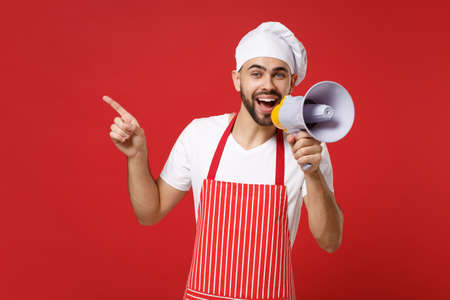 Cheerful bearded chef cook or baker man in striped apron t-shirt toque chefs hat isolated on red background. Cooking food concept. Mock up copy space. Scream in megaphone, pointing index finger aside.の写真素材